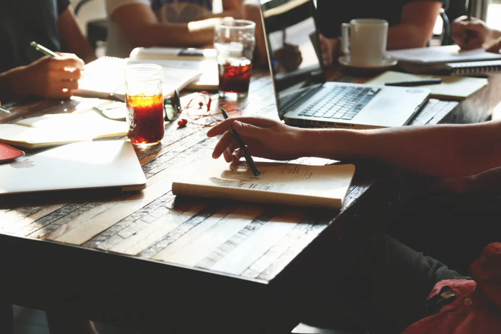 A high-angle, close-up shot of a rustic wooden table where people are working. A person’s hand is in focus, holding a pen over an open notebook filled with handwritten notes. Around the table are laptops, glasses of iced coffee, white mugs, and other notebooks, captured in warm, natural sunlight that creates a productive, collaborative atmosphere.